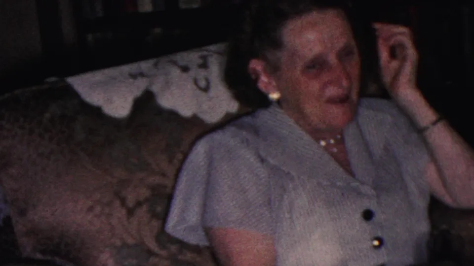 A woman in mid-century dress sits forward on a patterned couch with her hand lifted beside her head in a softly exposed Cleveland home-movie frame.