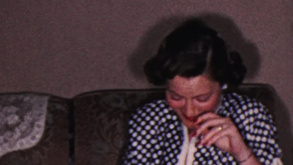A second woman appears seated on a couch in the same private interior, wearing a dark polka-dot garment and looking downward with her hand near her mouth.