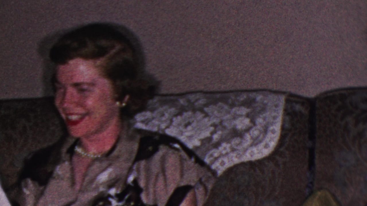 1953 Cleveland home-movie frame showing an adult woman with curled hair, a pearl necklace and a dark-trimmed blouse laughing on a patterned couch.