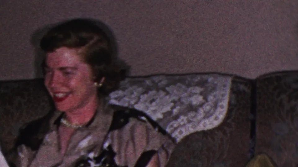 1953 Cleveland home-movie frame showing an adult woman with curled hair, a pearl necklace and a dark-trimmed blouse laughing on a patterned couch.