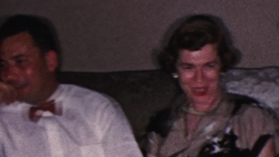 1953 Cleveland home-movie frame showing a man in a white dress shirt and red bow tie seated near a woman with curled hair on a patterned living-room couch.
