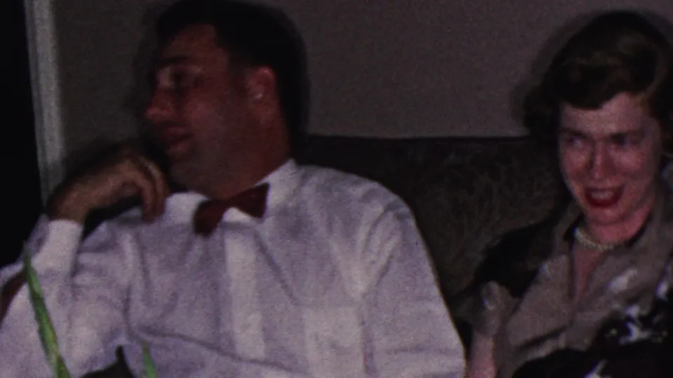 1953 Cleveland home-movie frame showing a man in a white shirt with a red bow tie turned toward a smiling woman seated beside him on a patterned couch.