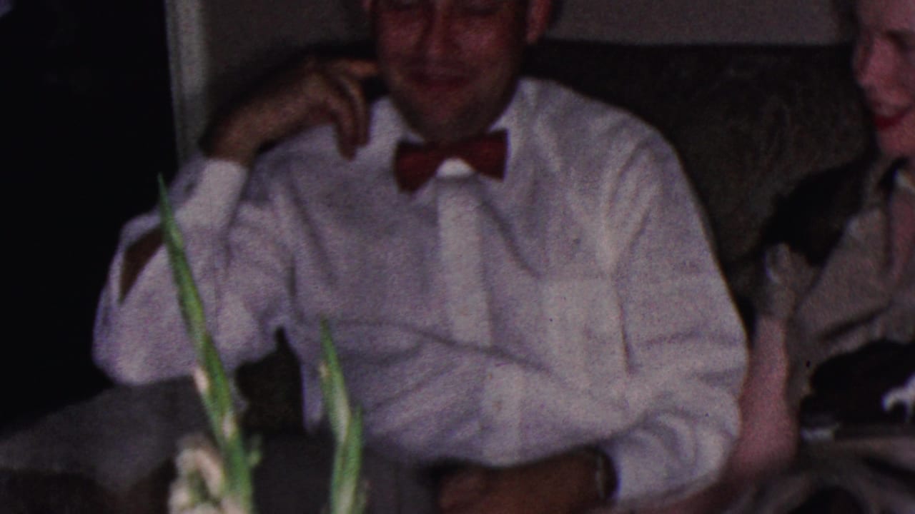 In a 1953 Cleveland home-movie frame, a man in a white dress shirt and red bow tie sits smiling near other adults in a softly lit private interior.
