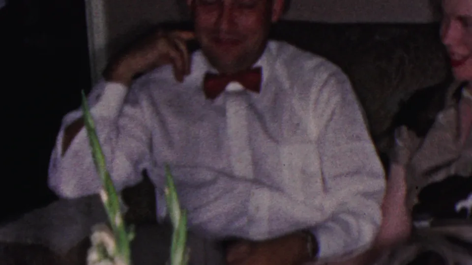 In a 1953 Cleveland home-movie frame, a man in a white dress shirt and red bow tie sits smiling near other adults in a softly lit private interior.