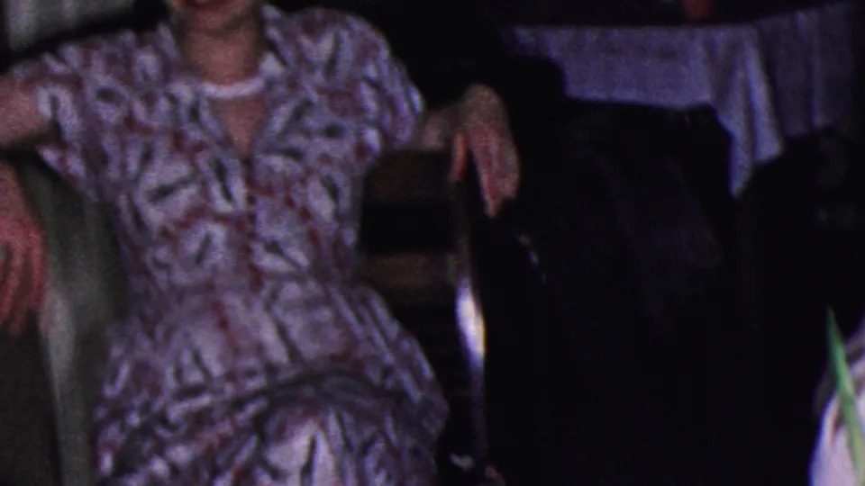 In this 1953 Cleveland frame, a woman in a patterned dress sits casually beside another adult, with upholstered furniture visible in the dim room.