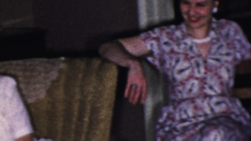 A woman in a patterned dress smiles from an upholstered seat in a 1953 Cleveland private interior, with the home-movie frame softly focused and grainy.