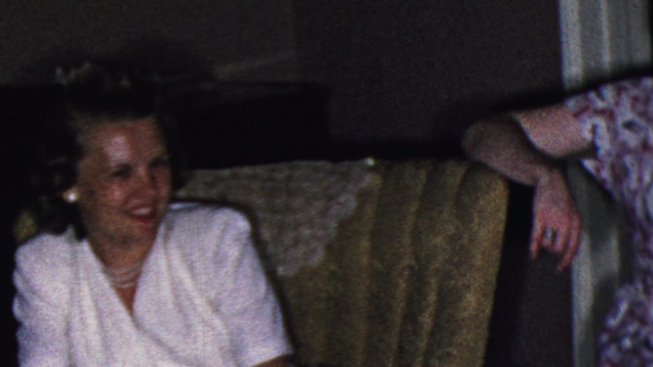 In a Cleveland, Ohio home-movie frame from the supplied 1953 metadata, a woman in a white dress smiles from a patterned sofa while other adults sit nearby in a dim living room.