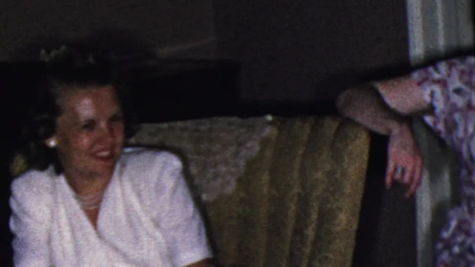 In a Cleveland, Ohio home-movie frame from the supplied 1953 metadata, a woman in a white dress smiles from a patterned sofa while other adults sit nearby in a dim living room.