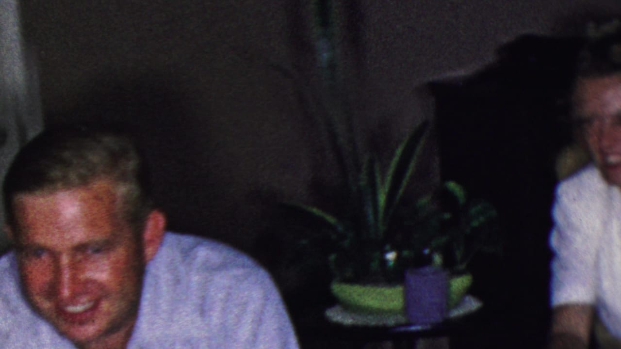 Man in a light shirt smiles near a sofa, plants, and a small table.