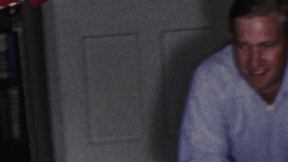 A man in a light shirt sits close to the camera inside the living room, with a closed interior door behind him and shelves or books visible along the side.