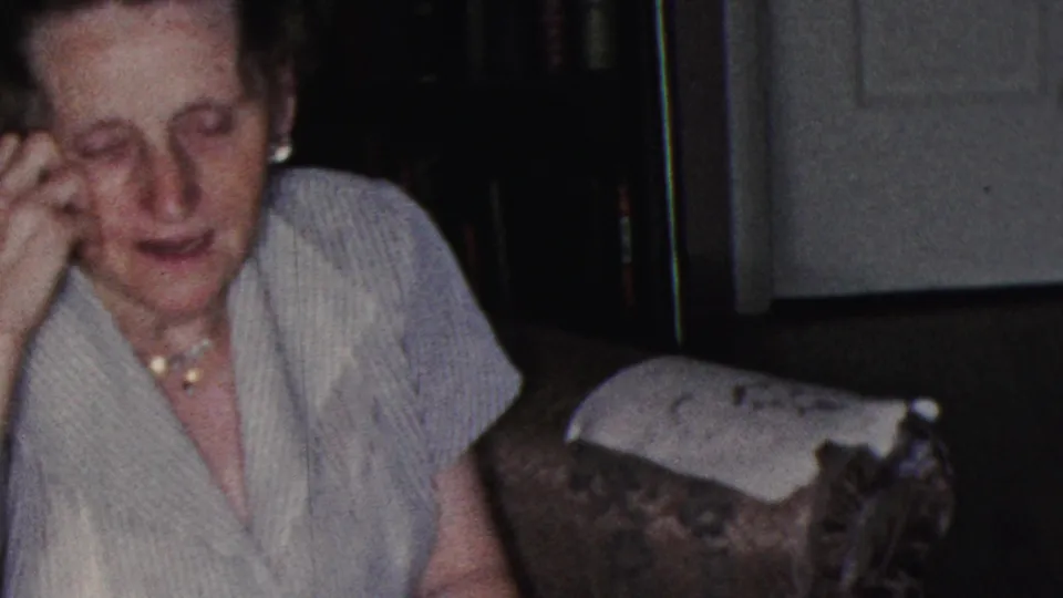 An older woman in a striped top sits beside a man in a Cleveland living room, with a dark bookshelf, white door, and upholstered chair visible in the background.