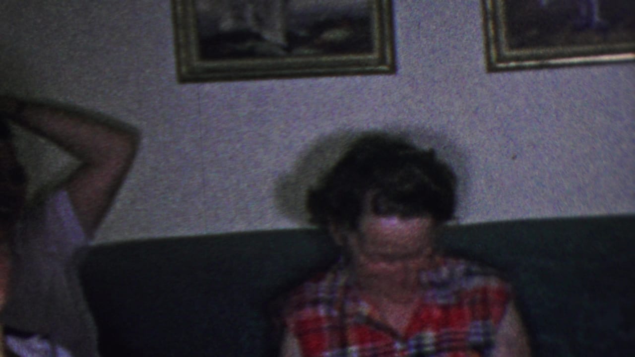 In this 1953 Cleveland home-movie frame, a woman in a colorful plaid sleeveless shirt sits on a dark couch beneath framed wall pictures in a private living room.