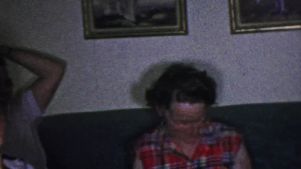 In this 1953 Cleveland home-movie frame, a woman in a colorful plaid sleeveless shirt sits on a dark couch beneath framed wall pictures in a private living room.