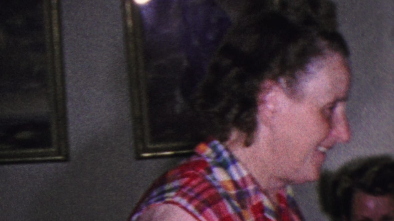 Smiling woman in a plaid sleeveless shirt seen in profile indoors.