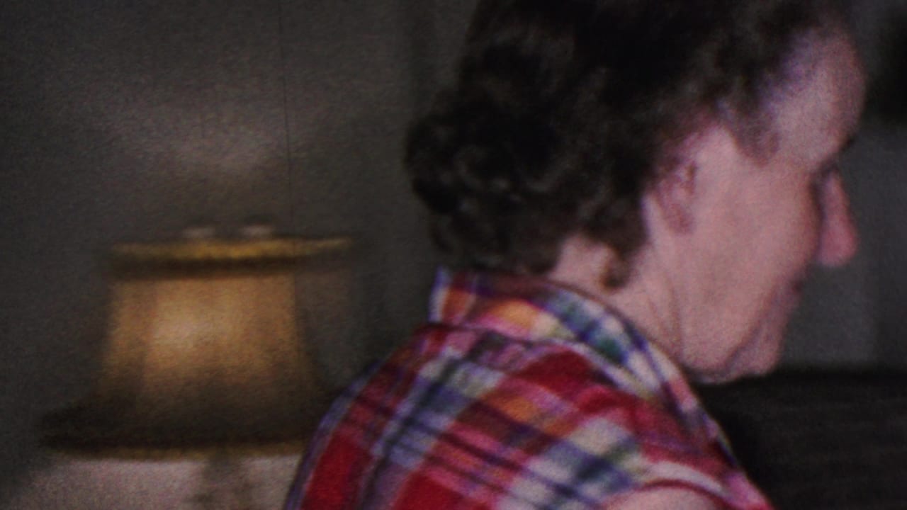 A close side view shows the woman in a plaid sleeveless shirt near a glowing table lamp, with the handheld 8mm frame emphasizing the private living-room setting.