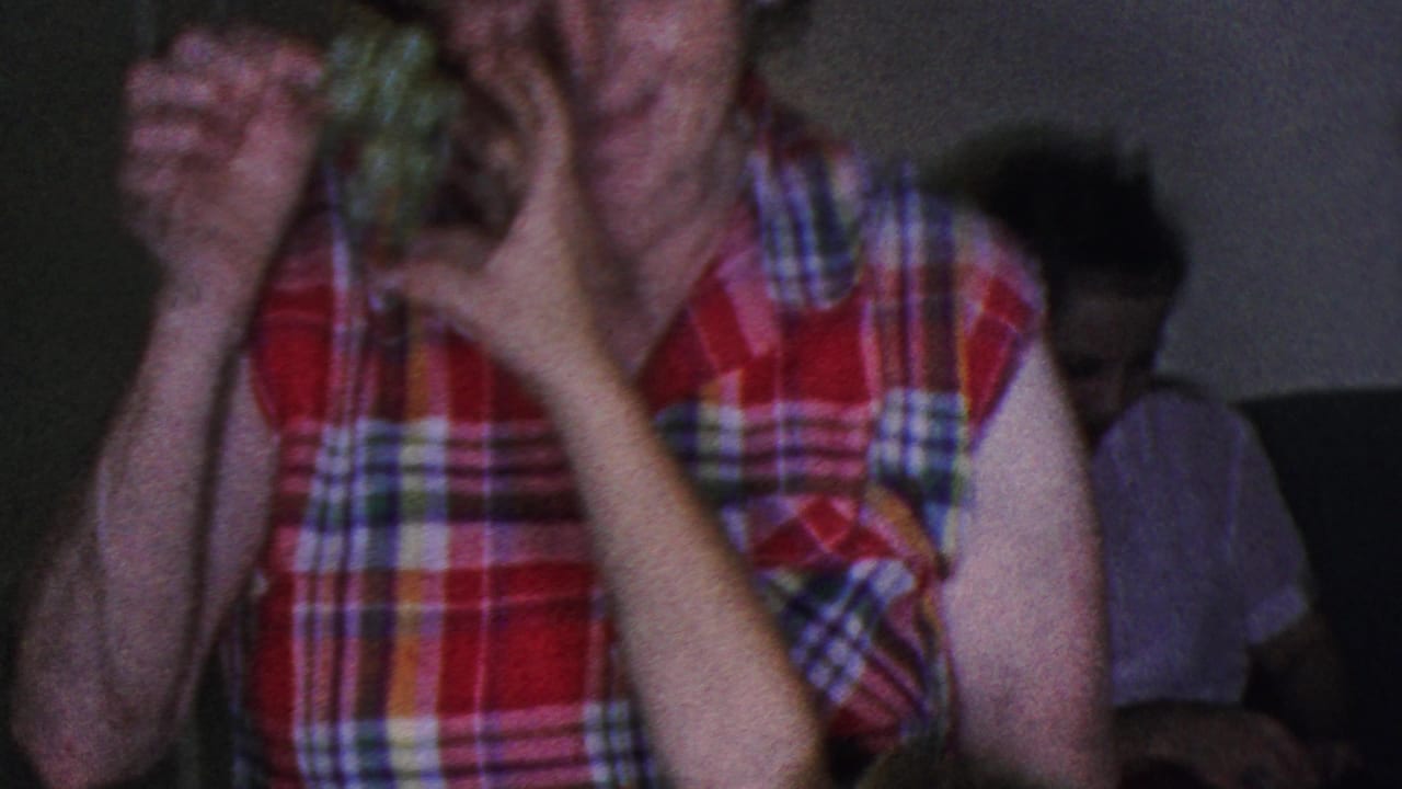 In 1953 Cleveland home-movie footage, a woman in a red plaid sleeveless blouse holds green grapes near her face while seated figures appear in the dim background.