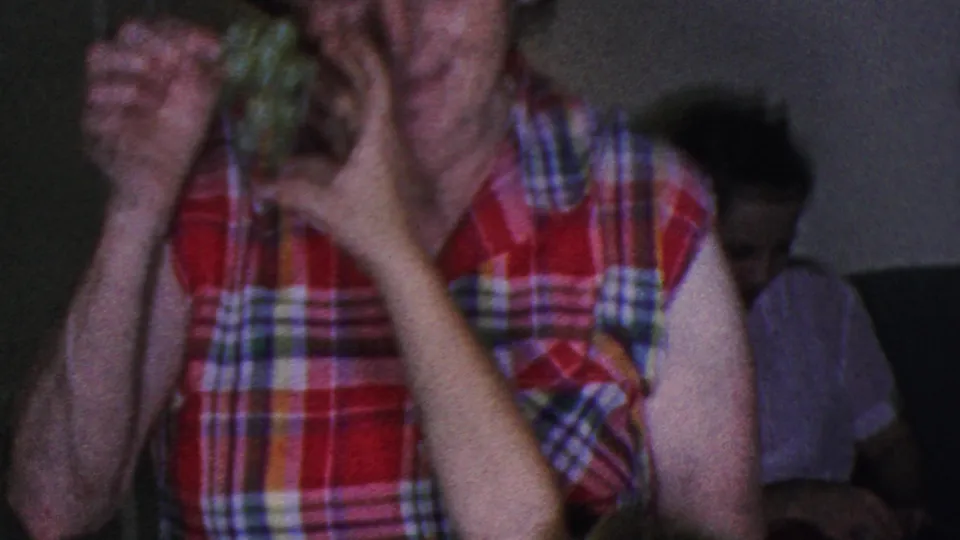 In 1953 Cleveland home-movie footage, a woman in a red plaid sleeveless blouse holds green grapes near her face while seated figures appear in the dim background.