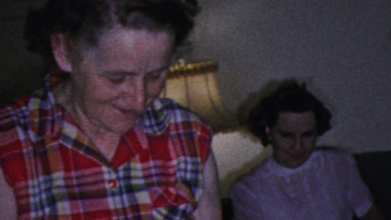A smiling woman in a red plaid sleeveless blouse looks downward in a Cleveland living room, with another woman seated in the background beside a shaded lamp.