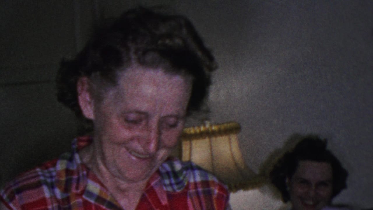 The 1953 Cleveland home-movie frame shows the woman in red plaid smiling downward while a seated woman and warm table lamp remain visible in the living room background.