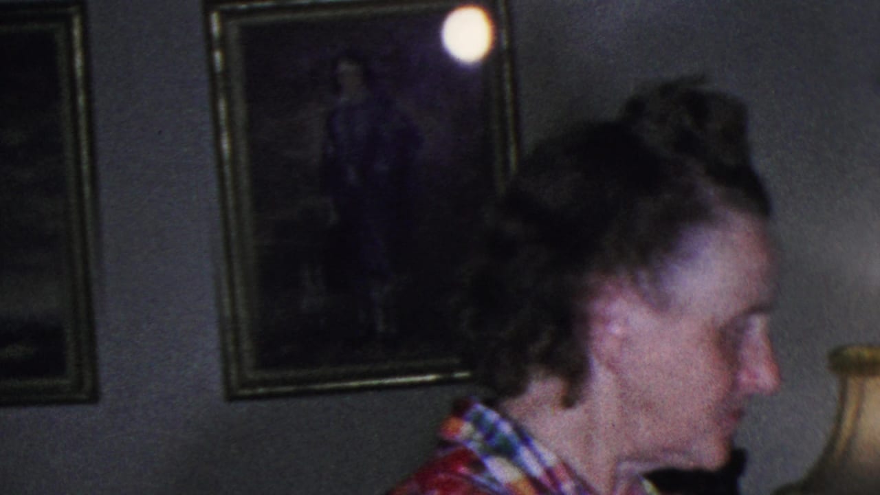 A woman in a red plaid sleeveless blouse turns in profile inside the Cleveland living room, with framed wall pictures and a shaded lamp visible behind her.