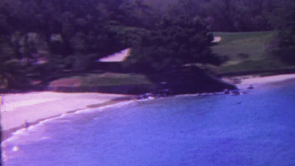 1962 Hawaii 8mm frame showing a quiet tropical beach with pale sand, blue surf, dense palms, rocks, and a distant person near the waterline.
