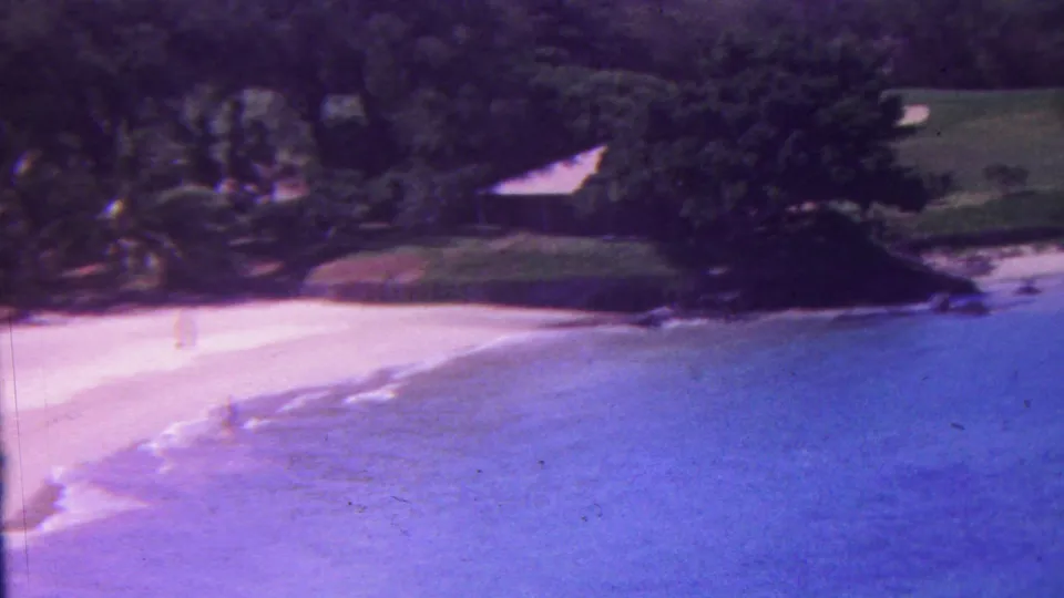 1962 Hawaii 8mm frame of a curving tropical shoreline with blue water, bright sand, dense vegetation, dark rocks, and a small structure partly hidden by trees.