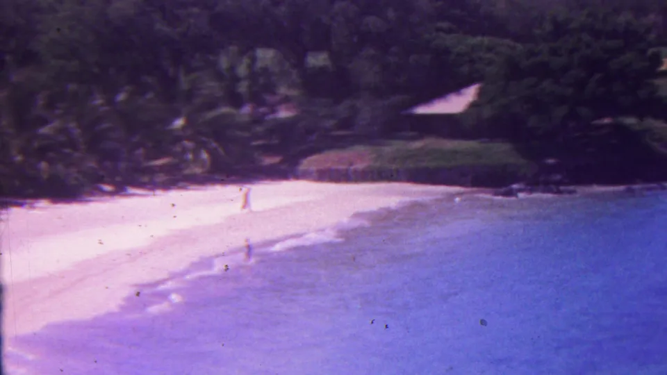 1962 Hawaii 8mm frame showing a bright palm-lined beach, a distant walker near the water's edge, blue surf, dark rocks, and a low structure set back in greenery.