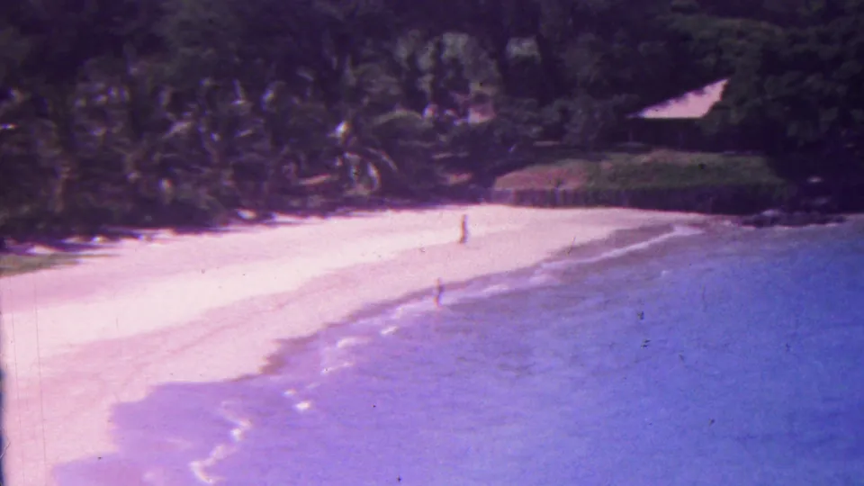 1962 Hawaii 8mm frame of a broad sandy shoreline with blue water, low waves, palms, dark rocks, and one distant figure standing or walking on the beach.