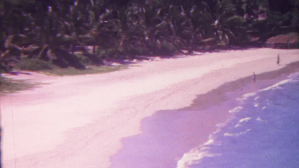 1962 Hawaii 8mm frame with a curving beach, pale sand, blue surf, palm vegetation, and small figures spaced along the shoreline.