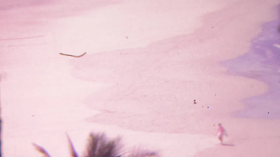 1962 Hawaii 8mm frame showing bright pink-toned sand, blue-purple surf, palm fronds in the foreground, and a distant person walking near the lower right edge.