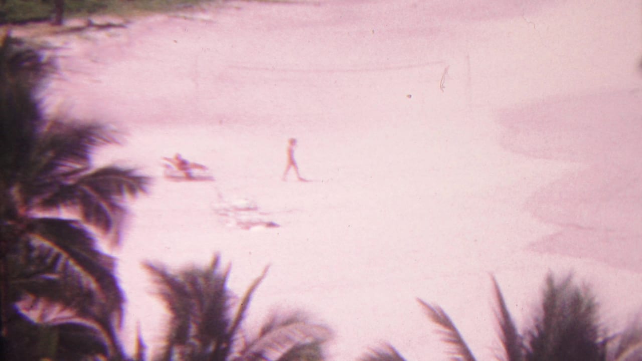 A pink-shifted 1962 Hawaii beach frame shows one person walking across open sand while another beachgoer rests near a chair below palm fronds.