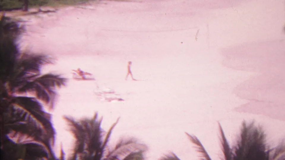 A pink-shifted 1962 Hawaii beach frame shows one person walking across open sand while another beachgoer rests near a chair below palm fronds.