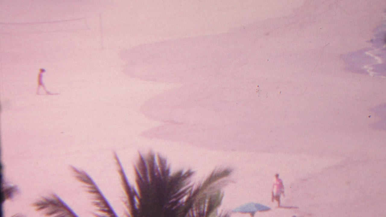 A 1962 Hawaii beach frame shows palm fronds in the foreground, a lone distant figure on sand, a small blue umbrella or shade, and surf entering at the right edge.