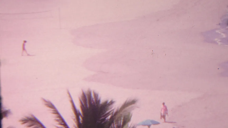 A 1962 Hawaii beach frame shows palm fronds in the foreground, a lone distant figure on sand, a small blue umbrella or shade, and surf entering at the right edge.
