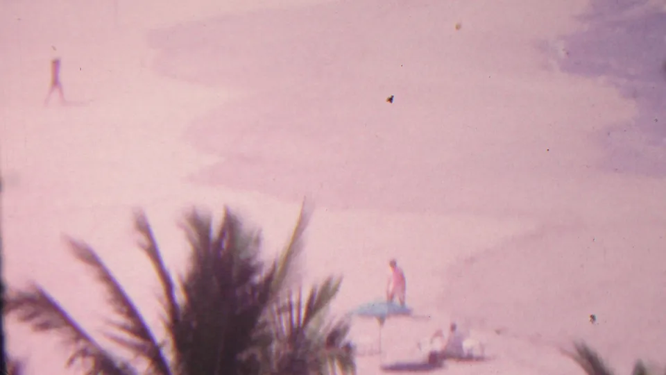 1962 Hawaii 8mm frame showing a high-angle view over palm fronds toward beachgoers, blue umbrellas, lounge chairs, and shallow shoreline water.