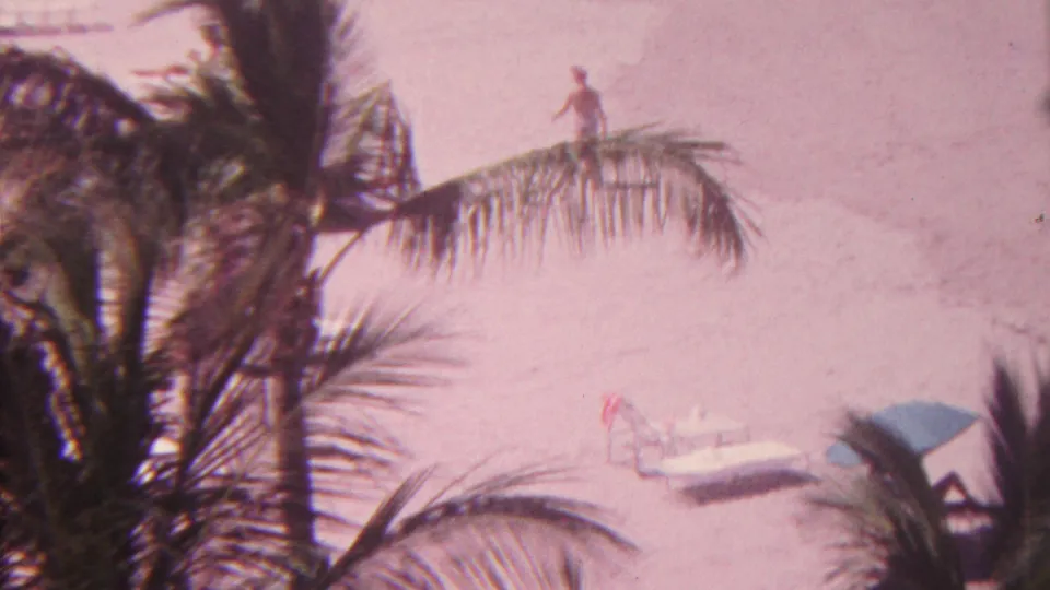 1962 Hawaii 8mm frame looking down through palm trees onto a pale sandy beach with lounge chairs, a blue umbrella, and a distant beachgoer near gentle surf.