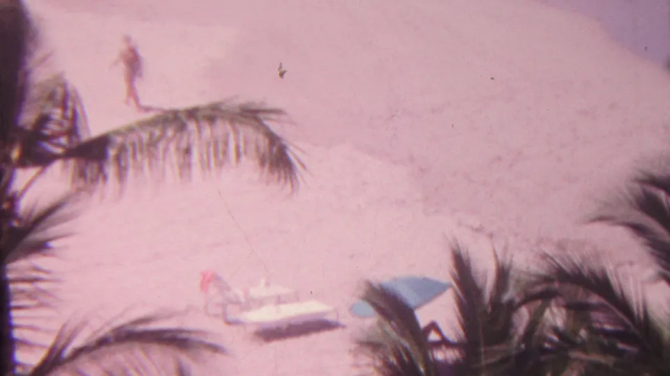 1962 Hawaii beach view with a distant person walking along pale sand, palm fronds crossing the frame, lounge chairs, a blue umbrella, and surf at the upper edge.