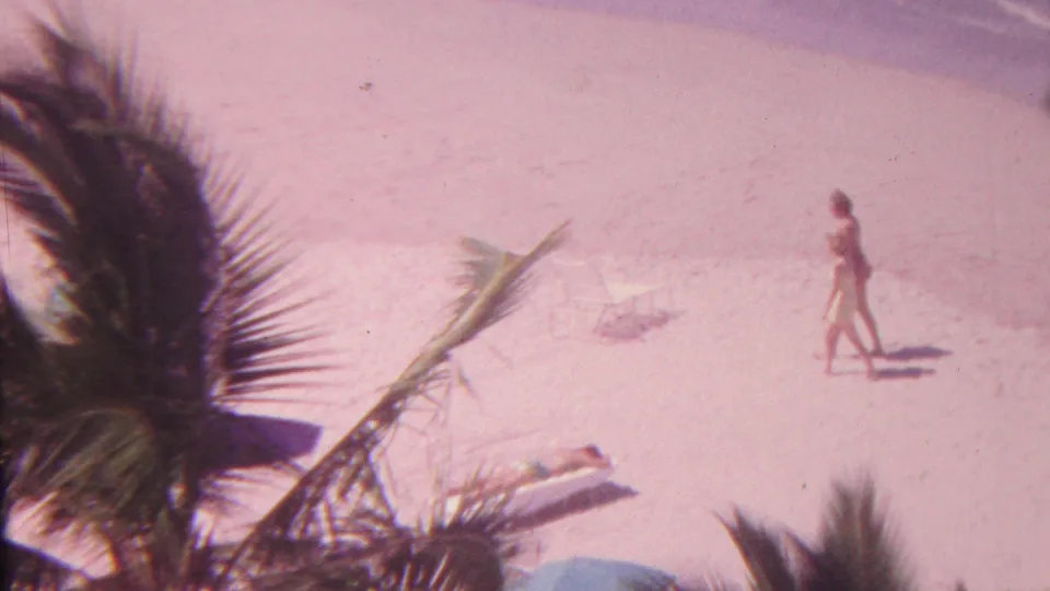 1962 Hawaii 8mm frame showing a beachgoer walking across sunlit sand below palm fronds, with lounge chairs, beach items, and ocean water nearby.
