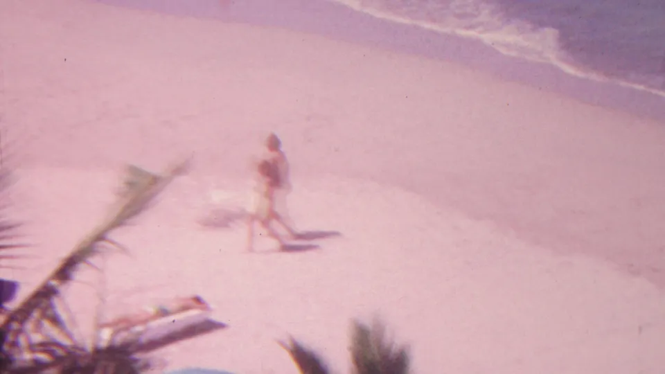 1962 Hawaii beach frame with a distant person walking beside the surf, palm fronds in the foreground, a lounge chair on the sand, and gentle waves at the top of frame.