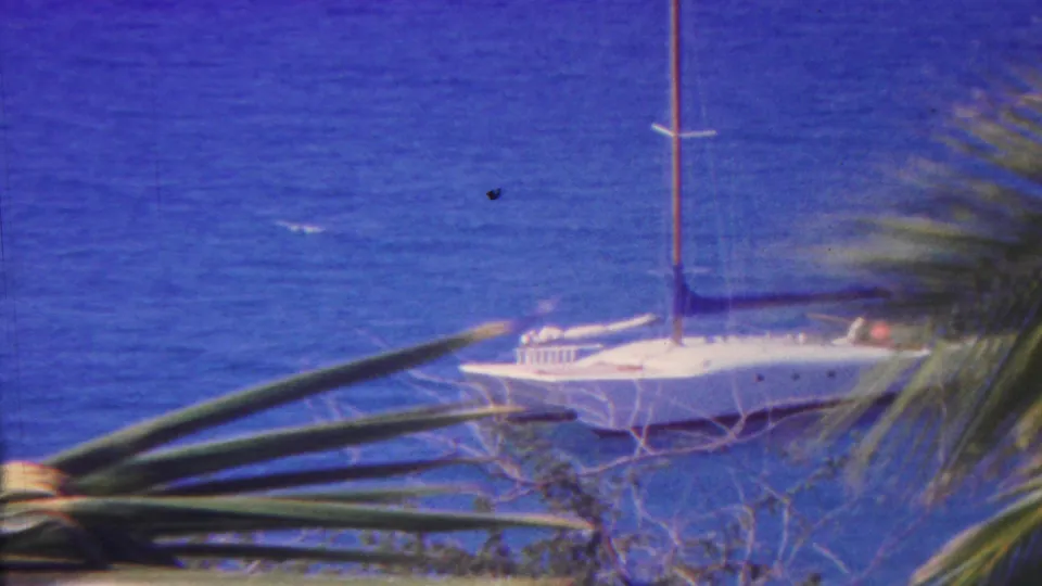 1962 Hawaii 8mm frame with a white anchored sailboat on blue ocean water, seen through palm fronds and sparse shoreline branches.