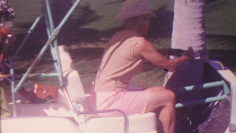 A man wearing light clothing and a wide-brimmed hat sits in a small open cart near a palm tree in the Hawaii home-movie scene.