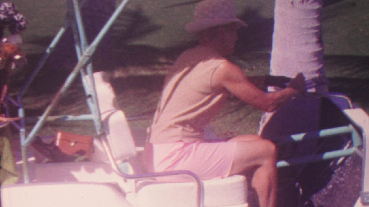 In Hawaii 1962 home-movie footage, a man in a light sleeveless shirt, pink shorts, and brimmed hat is seen from the side near the steering wheel of a white golf cart under bright sun.