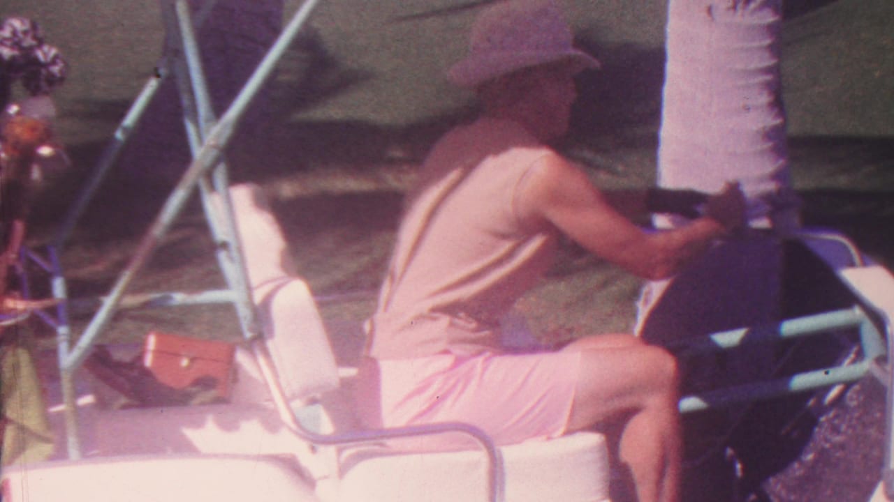 Side view of a man steering a white golf cart near palm trees and shaded grass.