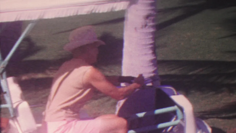 A soft 8mm frame from the Hawaii golf-course scene shows the man seated beside the white cart, wearing shorts and a brimmed hat, with a palm trunk and shaded turf in the background.