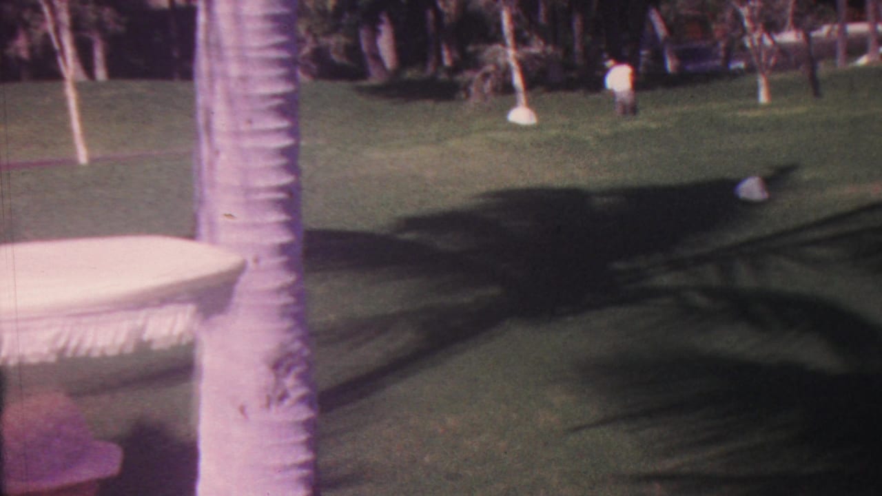 In 1962 Hawaii home-movie footage, a woman in a pale sleeveless outfit and wide-brimmed hat walks away across a green lawn with palm trunks, shadows, and a white fringed table nearby.