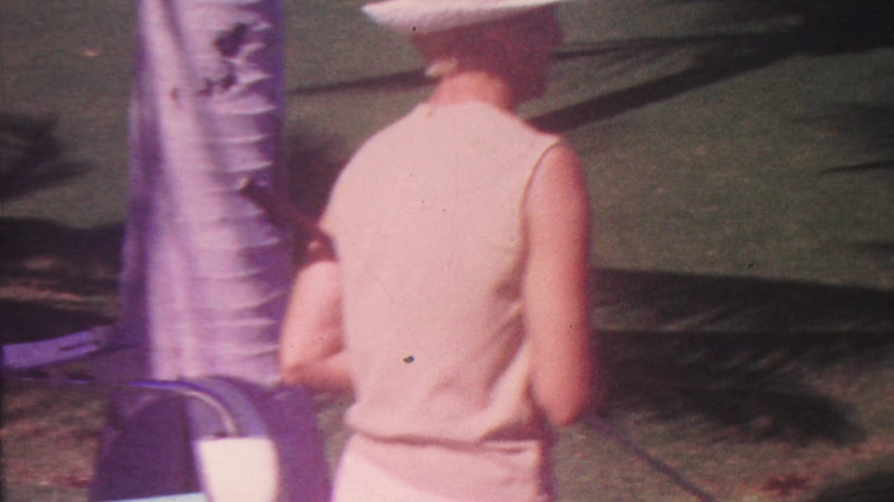 In the 1962 Hawaii clip, the woman continues across the lawn past a palm trunk, with strong palm shadows on the grass and a croquet ball visible ahead.