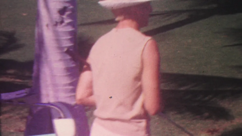 In the 1962 Hawaii clip, the woman continues across the lawn past a palm trunk, with strong palm shadows on the grass and a croquet ball visible ahead.