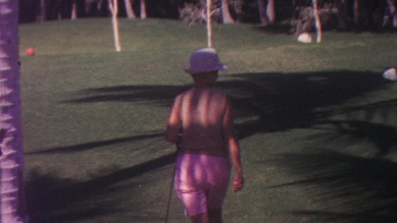 In Hawaii 1962 home-movie footage, a woman seen from behind walks across a manicured green lawn with a croquet mallet, with palm shadows and foliage in the background.