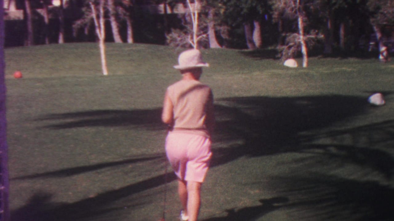 Woman in a hat walks away across a sunny lawn toward a red croquet ball.