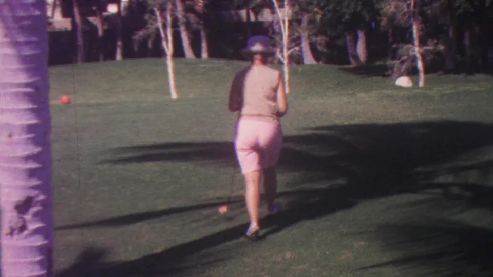 The 1962 Hawaii home-movie frame shows the woman from behind as she approaches a red croquet ball on a trimmed lawn, with a palm trunk at left and wooded greenery behind.